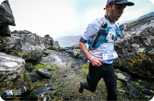 A man wearing a race bib runs on a rocky trail in a mountainous area, under cloudy skies.