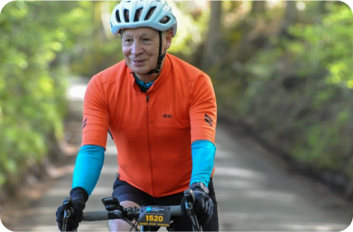 A cyclist wearing an orange jacket, blue sleeves, and a white helmet rides on a shaded road with greenery in the background.