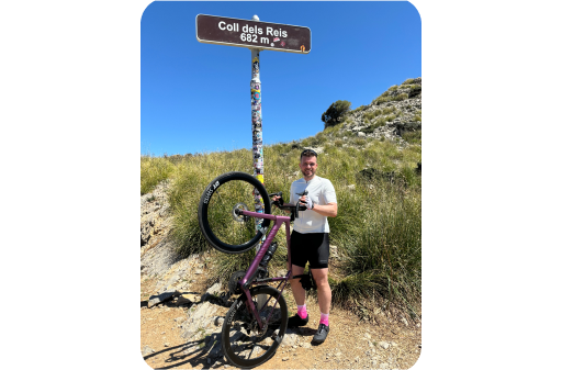 A cyclist stands beside a bike held upright on its rear wheel under a sign reading "Coll dels Reis 682 m"—a victorious moment on a sunny, grassy hillside that could inspire anyone to embrace adventure.