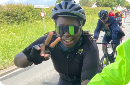 A cyclist wearing sunglasses and black gear smiles and makes a peace sign while riding on the road with other cyclists, promoting PDA (Pathological Demand Avoidance) awareness in the vibrant cycling community.