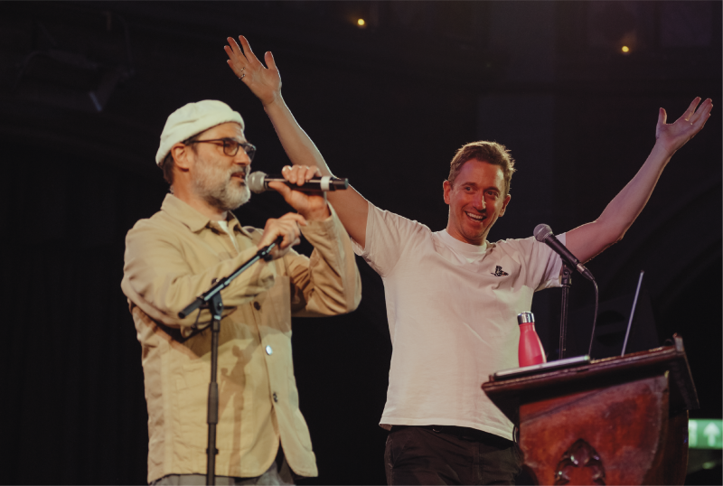Two men stand on stage at microphones; one speaks while the other raises his arms and smiles. A red bottle sits on the podium in front of them.