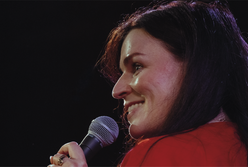 A woman with long dark hair smiles while holding a microphone, wearing a red top, against a dark background.