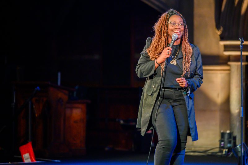 A woman with long braided hair, wearing glasses and dressed in black, stands on stage holding a microphone and smiling.