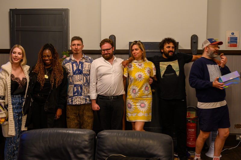 Seven adults stand in a row indoors, some smiling, dressed in casual and patterned clothing, with a fire extinguisher and gray chairs visible in the background.