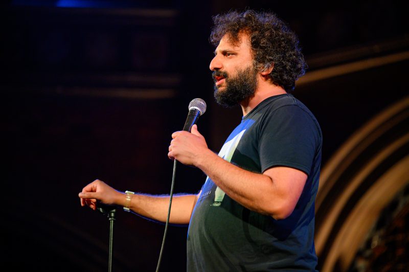 A man with curly hair and a beard holds a microphone and speaks on stage, wearing a black t-shirt, against a dark, blurred background.