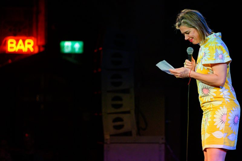 A woman in a yellow floral dress stands on stage holding a microphone and reading from a piece of paper, with a neon "BAR" sign visible in the background.