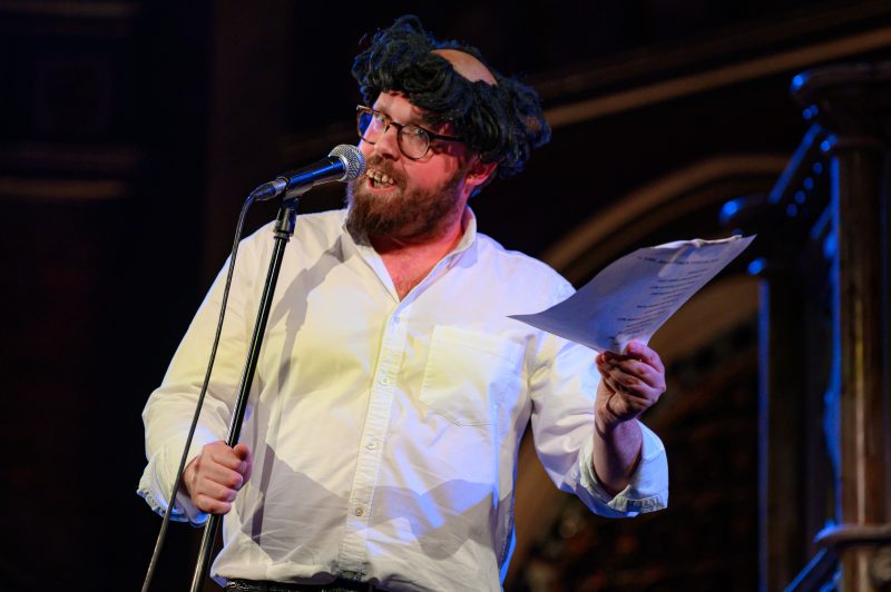 A man in a white shirt and glasses stands at a microphone, holding papers and wearing a humorous fake wig on stage.