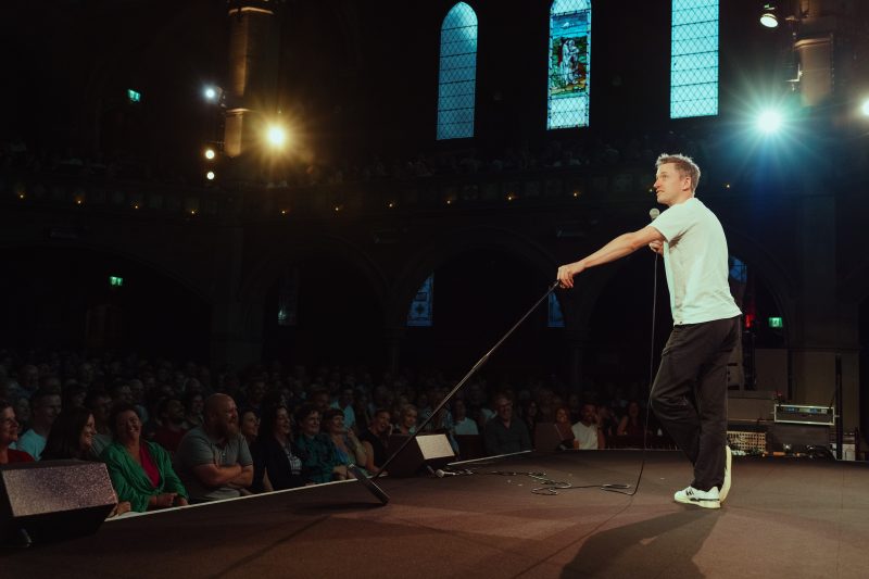A man in a white t-shirt and dark pants performs on stage with a microphone in front of a seated audience in a dimly lit venue with arched windows.