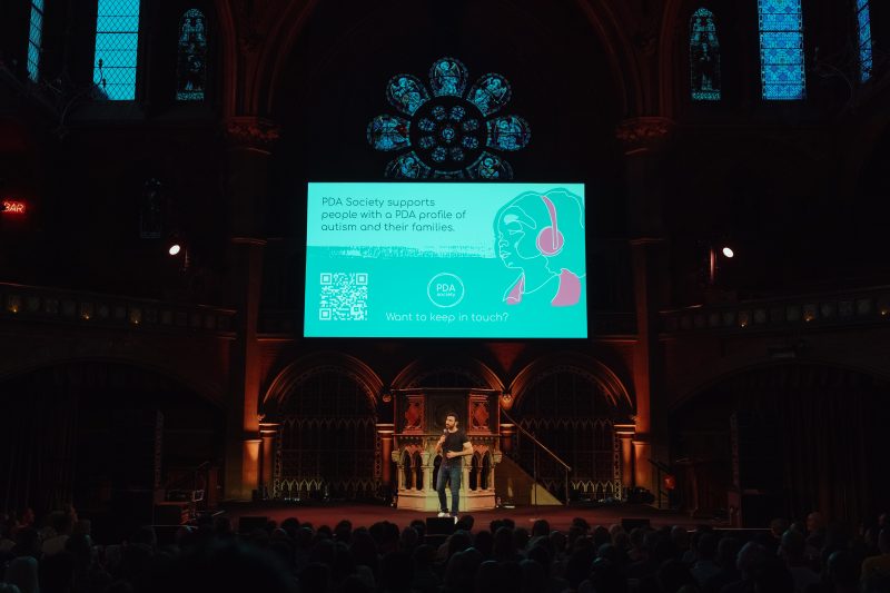 A speaker stands on stage in a dimly lit hall, presenting in front of a screen about PDA Society and autism to an audience.