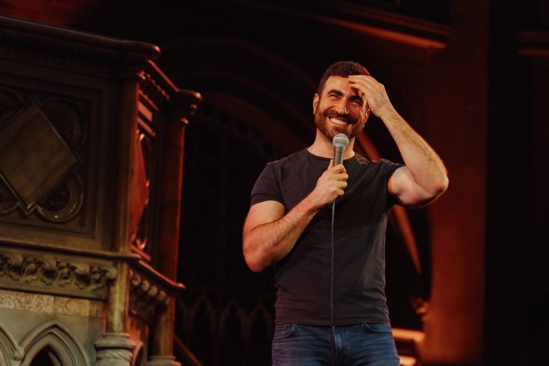 A man with a beard smiles while holding a microphone on a dimly lit stage with ornate architectural details in the background.