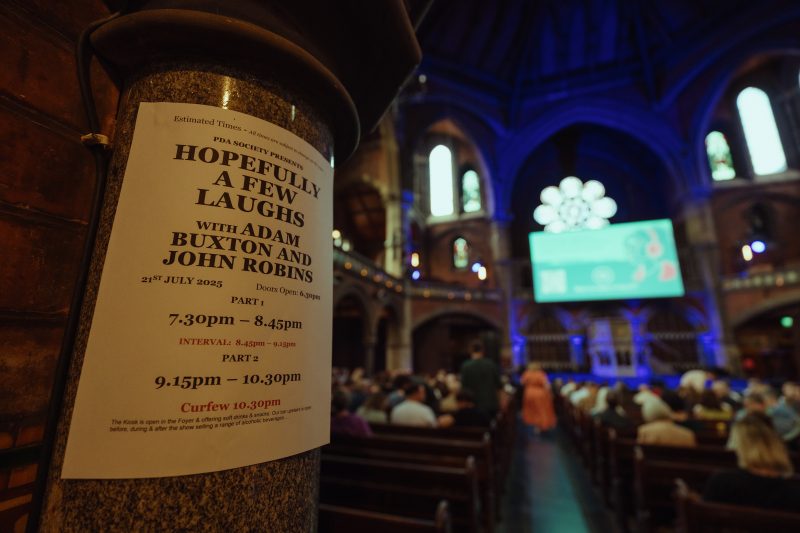 Poster for a comedy event featuring Adam Buxton and John Robins inside a church; audience is seated facing a screen at the front of the venue.