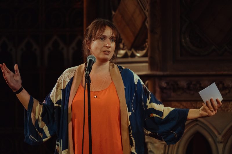 A woman stands in front of a microphone, gesturing with open arms, wearing a patterned robe over an orange top, and holding a notecard in one hand.