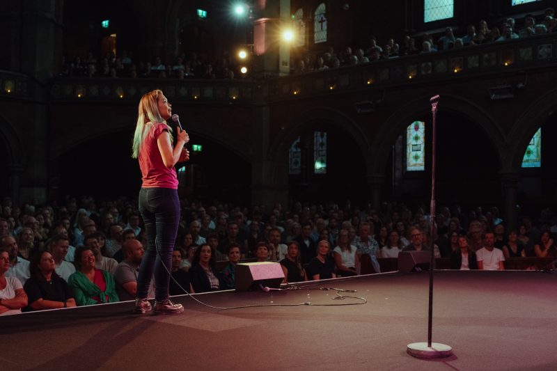 A person with long blonde hair stands on stage holding a microphone, facing a large seated audience in a dimly lit theater with arched windows.