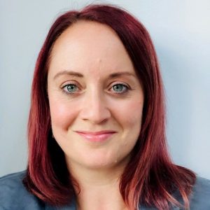 A woman with shoulder-length reddish-brown hair and blue eyes smiles at the camera, wearing a grey blazer in front of a plain light-coloured background, introducing the new Chief Executive of the PDA Society.