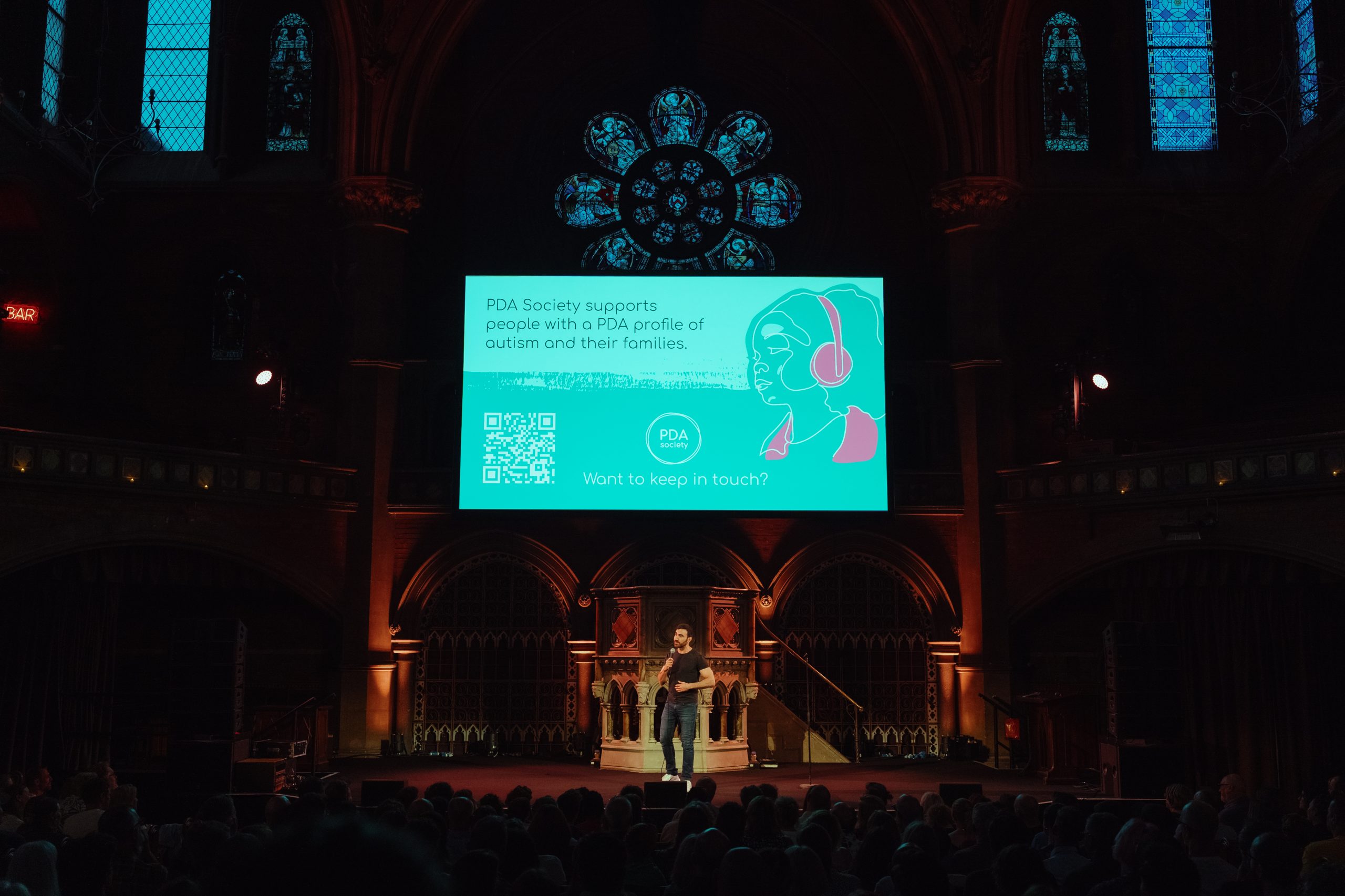 A speaker stands on stage in a dimly lit hall, presenting in front of a screen about PDA Society and autism to an audience.