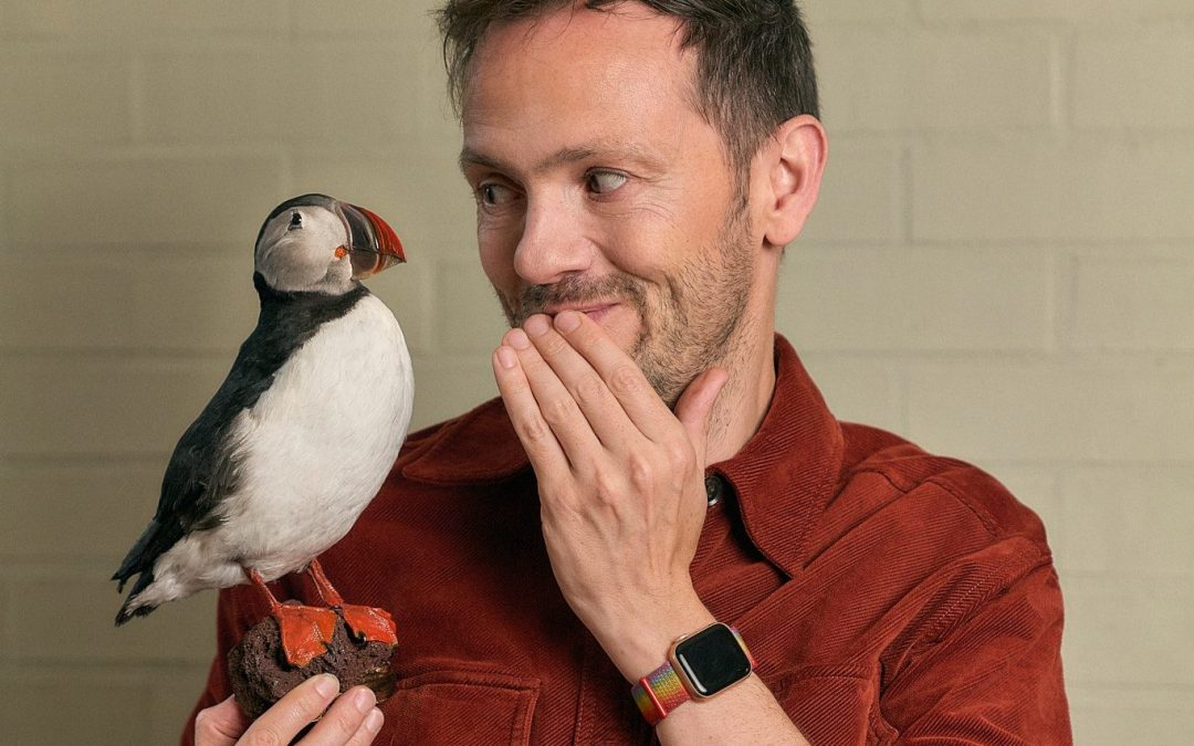 Stuart Laws, in a rust-colored shirt, holds a puffin model and smiles—covering his mouth with one hand—as if mid-chat, standing in front of a light brick wall.