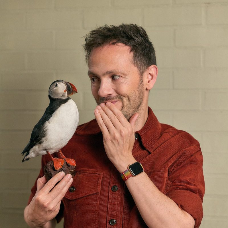 Stuart Laws, in a rust-colored shirt, holds a puffin and covers his mouth with one hand as if caught mid-interview, standing against a beige brick wall.